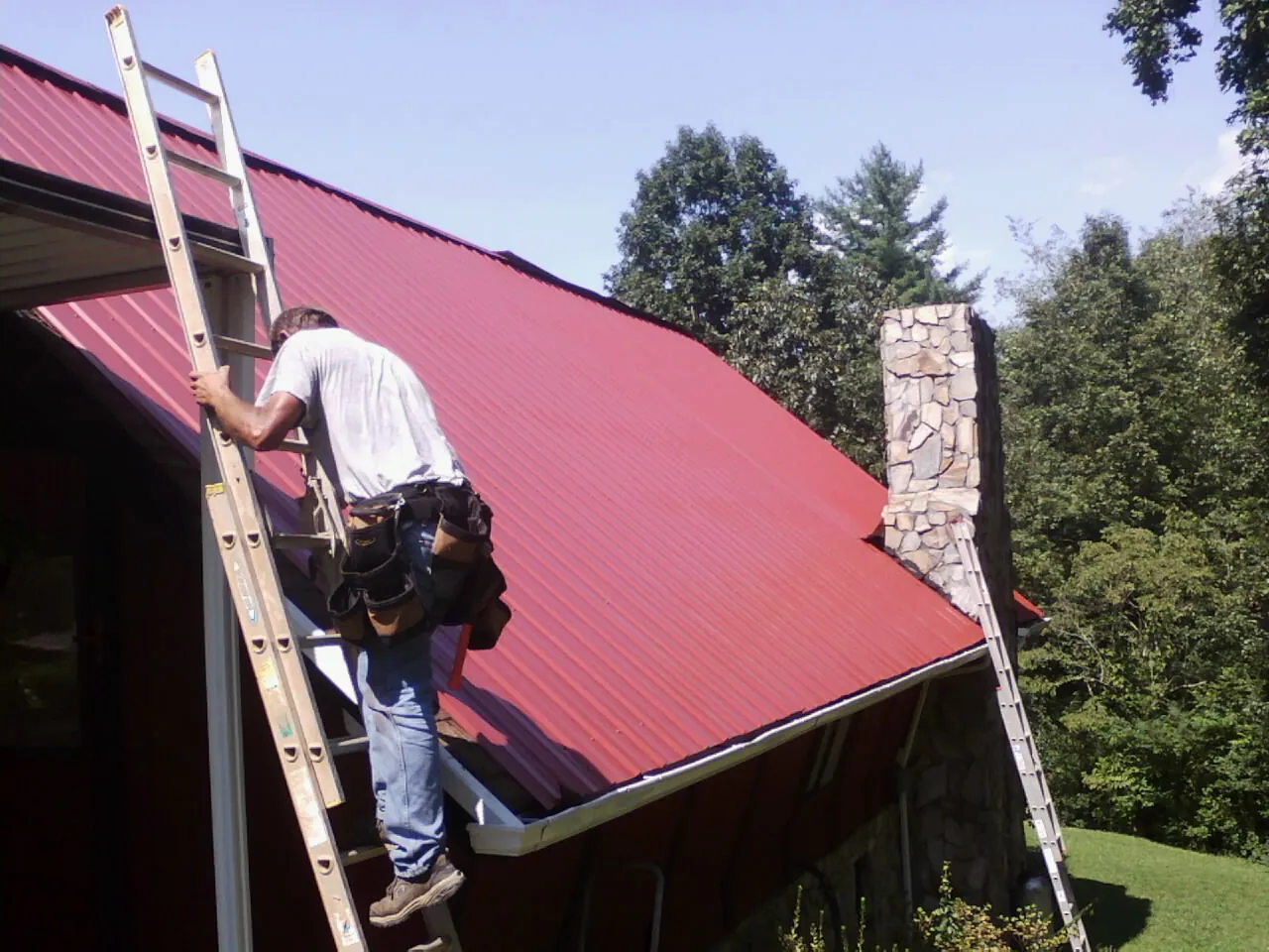 Roofer on a ladder beside a red metal roof and stone chimney — Soffit & Fascia Repair by Roofing Brothers in Rio Grande